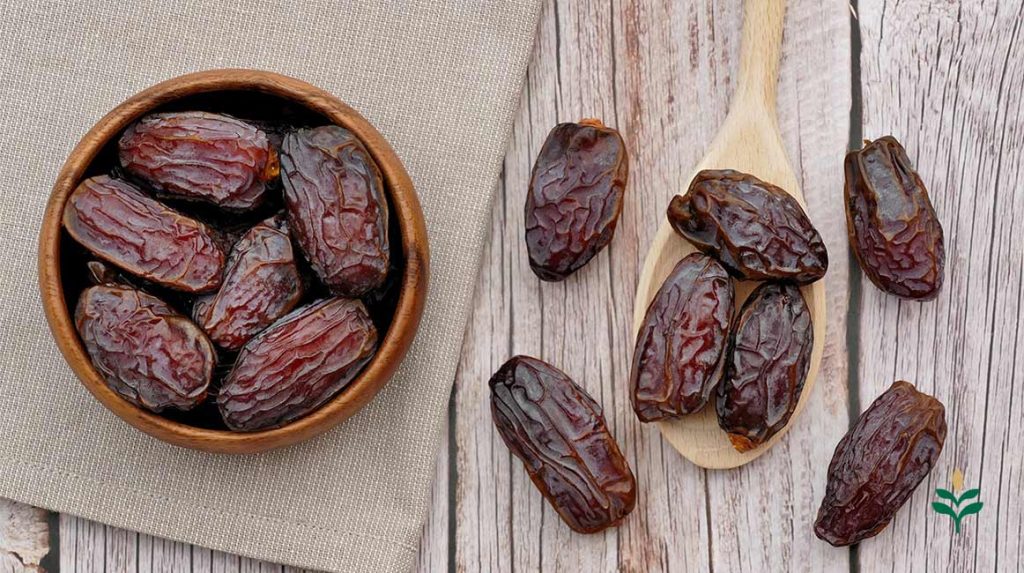 یک کاسه کوچک چوبی حاوی دانه های پیارم پرفیرو روی پارچه کتان خاکی رنگ و چند دانه خرما در یک قاشق چوبی کنار آن