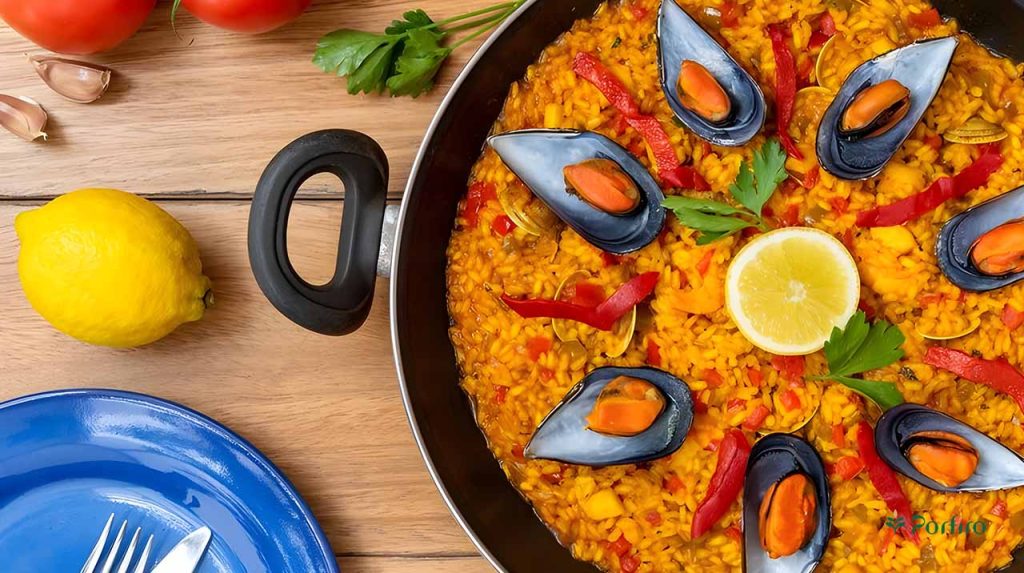 The side containing the paella is on a wooden table with a yellow lemon on it and a blue clay plate with a pair of knives and forks on the bottom left of the image.