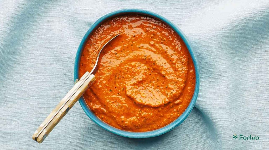 On a pale blue cloth background, in the middle of the image is a blue earthenware bowl containing romesco and a spoon.
