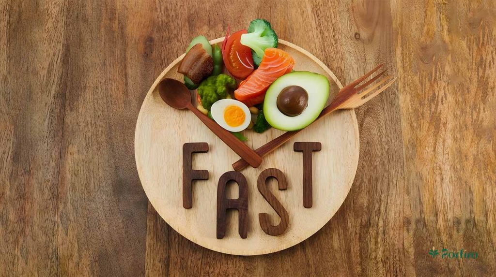 A wooden plate containing healthy nutrients and a pair of wooden spoons and forks with the word "fasting" written on them in wooden letters.