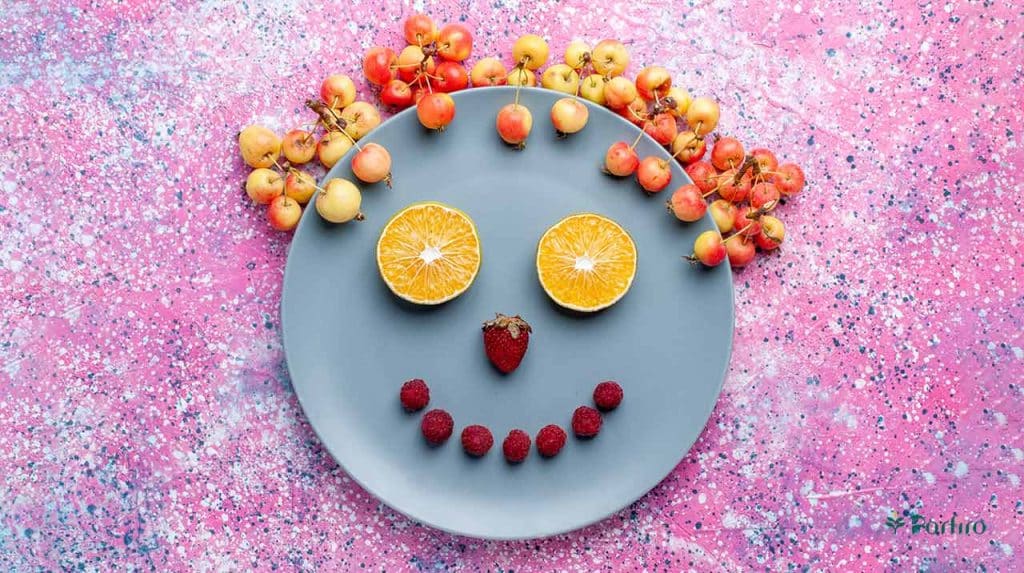 On a pink background is a plate with a smiley face inside, along with two orange halves, a few strawberries, and some cherries, encouraging conscious eating.