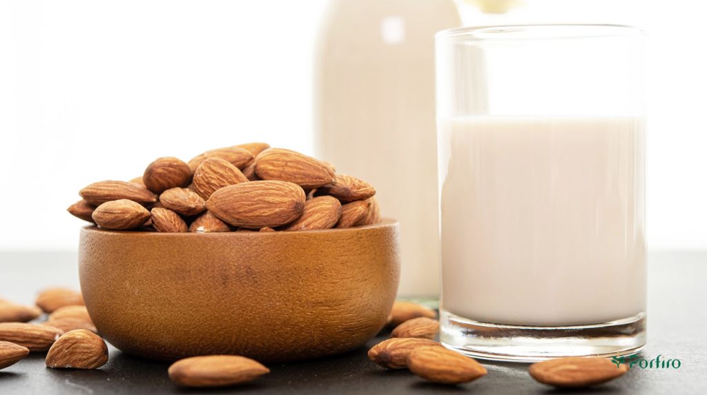 A bowl of almond kernels, with several of them on the table, and a glass of almond milk next to it.