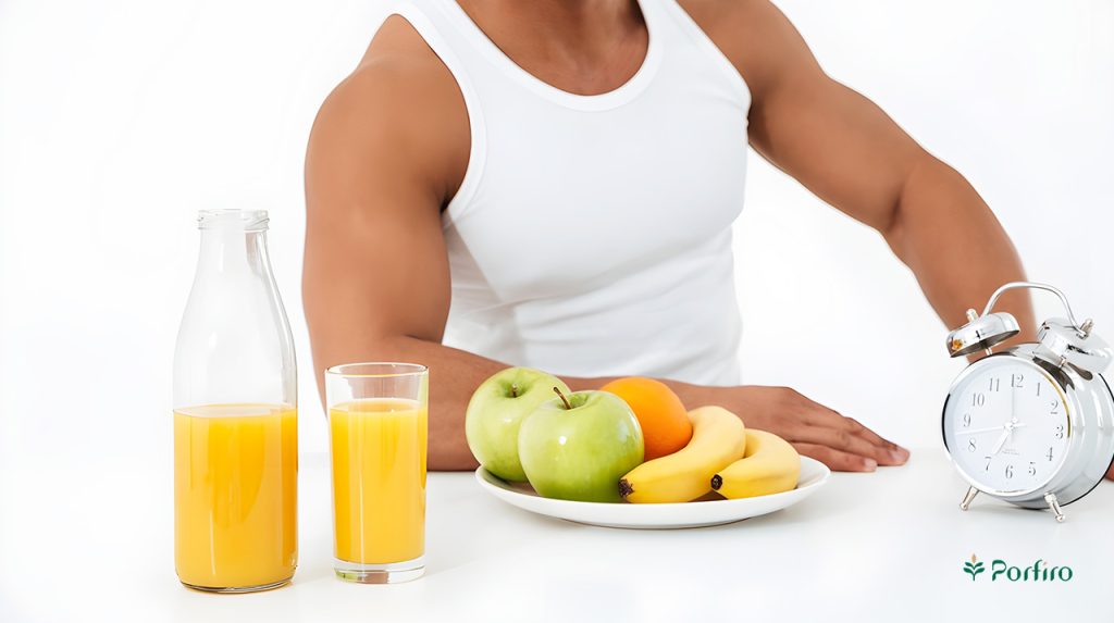The torso of an athletic man with well-muscled muscles, with a bowl of calorie-rich foods, such as bananas, apples, and oranges, on the table in front of him, along with a desk clock and a pitcher and glass of fresh orange juice.