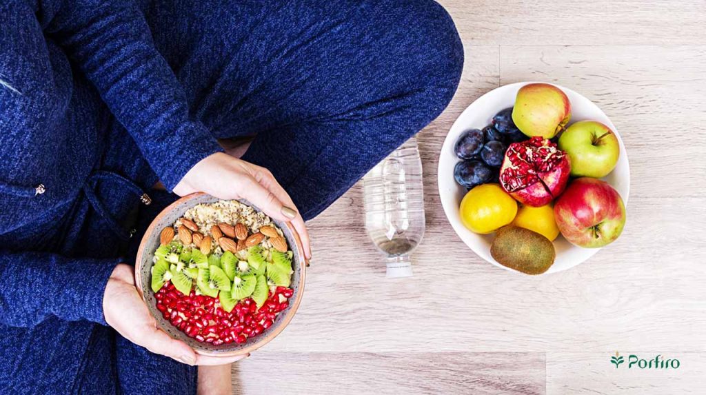 The lower torso of an athlete in sportswear with a container of suitable food in her hands and a container of fruit on the table in front of her