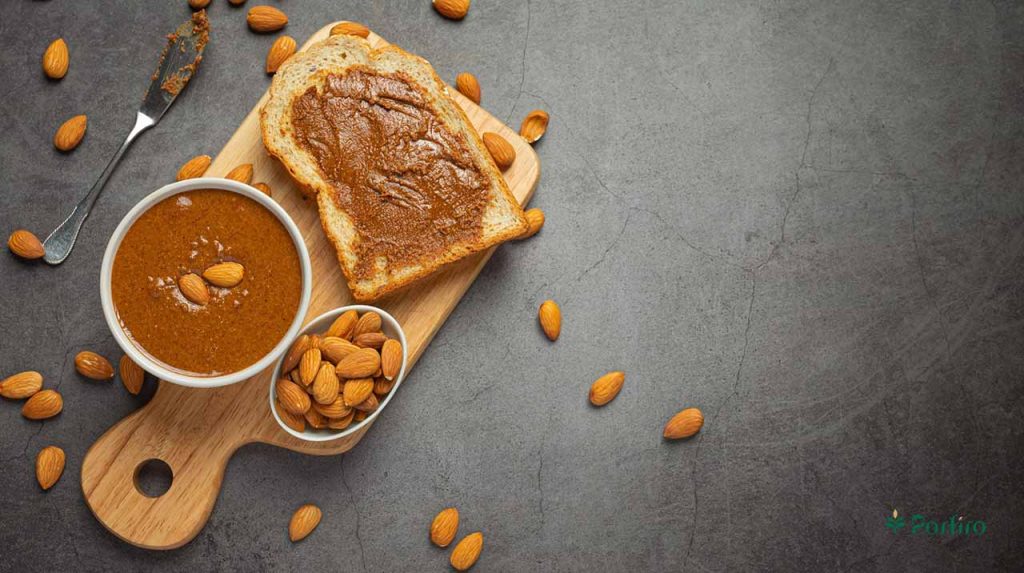A few almond kernels in one container, some almond butter in another container, and a slice of toast with almond butter spread on it, with a knife next to it.
