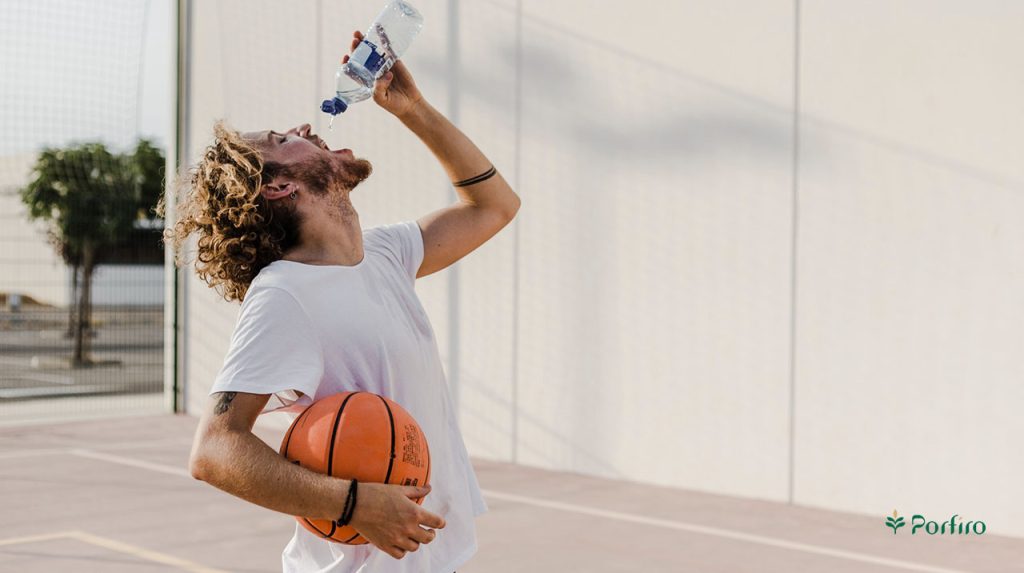 A male basketball player in a white t-shirt and curly hair is drinking water from a bottle while holding a basketball under his arm.