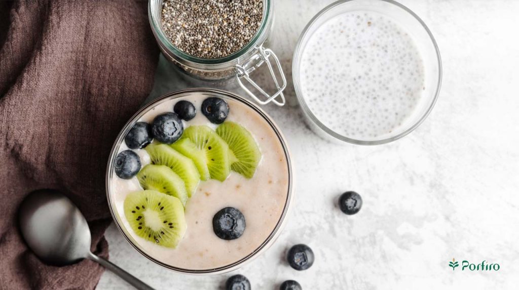A bowl full of dessert containing chia seeds, milk, blueberries, kiwi with a spoon next to it