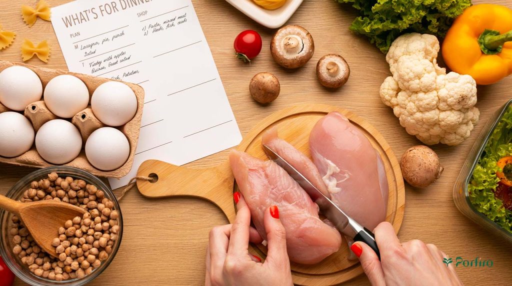 Woman's hands with red nail polish cutting chicken breast on a wooden kitchen board, surrounded by food items such as mushrooms, cabbage, eggs, peas and various bell peppers on the table