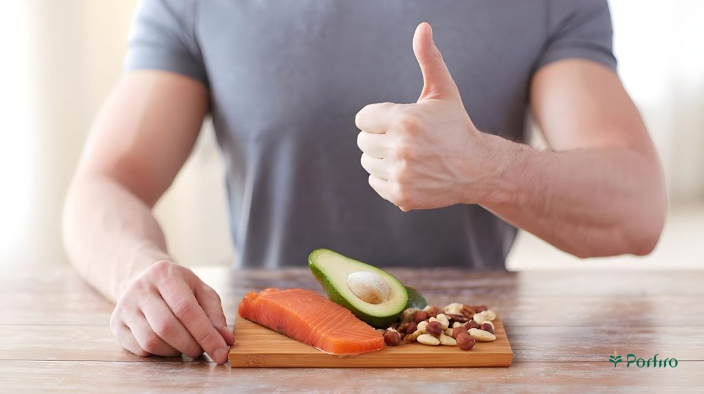 A male torso is showing a like sign with his hand and in front of him is a bowl containing various nuts, avocado, and a slice of salmon.