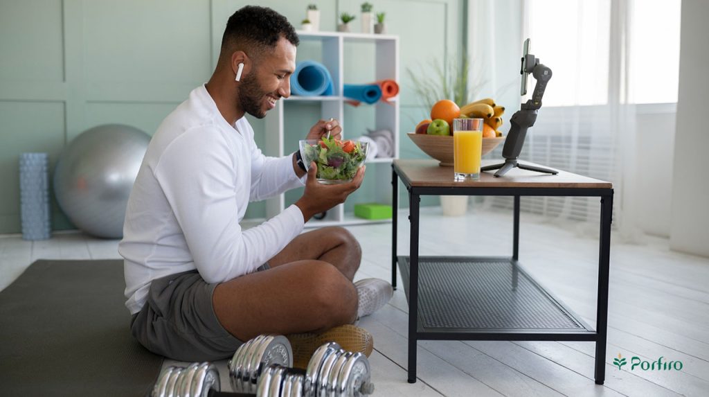 A male athlete is sitting on the floor in a gym and eating. On the table in front of him is a mobile phone with a stand, a fruit bowl, and a glass of juice.