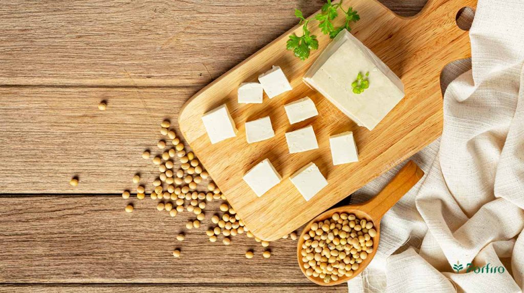 A tofu mold on a wooden worktop, with cubed slices of it, and a wooden spoon full of soybeans next to it.