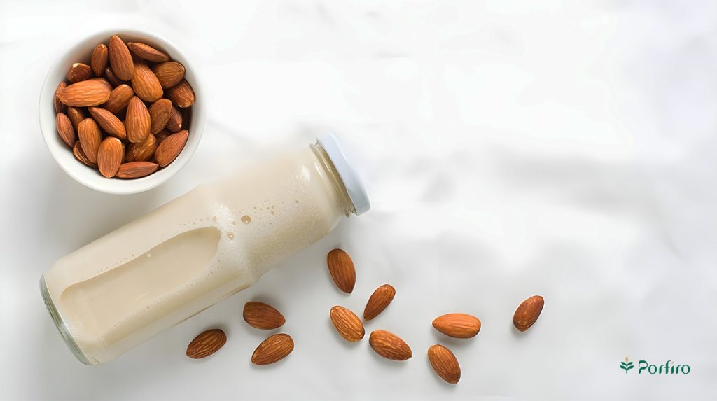 A glass bottle containing almond milk, with its lid closed, is placed horizontally on the table, with several almond kernels next to it, and a bowl containing almond kernels can also be seen to its left.