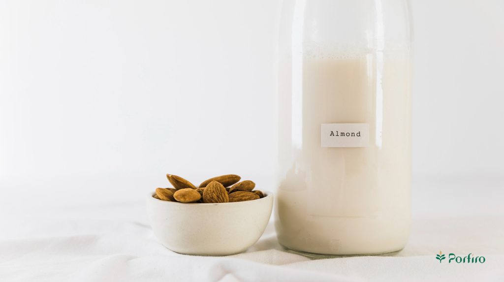 یک ظرف شیشه ای پر از شیر بادام و چنددانه مغز بادام در یک ظرف کوچک در کنار آن