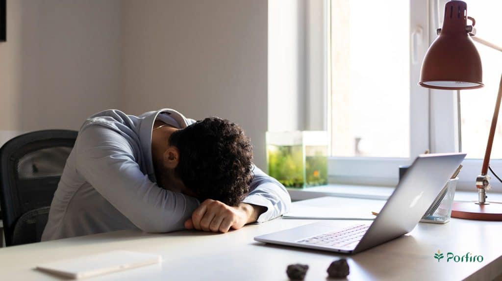 مردی پشت میز نشسته است و با لپ تاپ روی به روی او سرش را روی دستانش قرار داده است