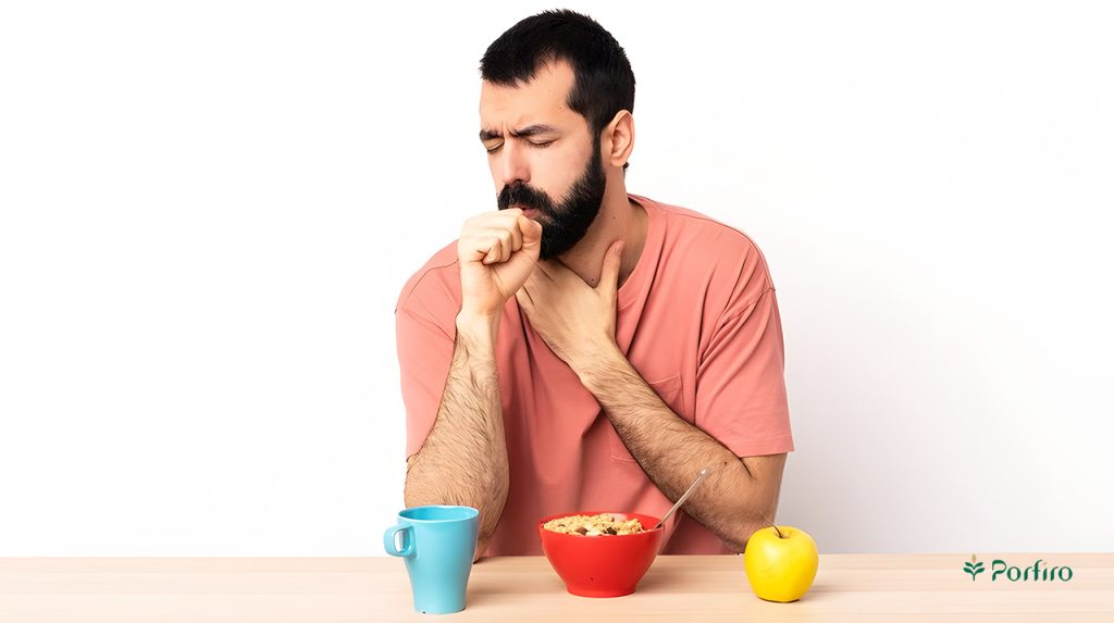 A man is sitting at a table coughing, and on the table in front of him is an apple, a glass of water, and a bowl of oatmeal. For the article on allergies or sensitivities