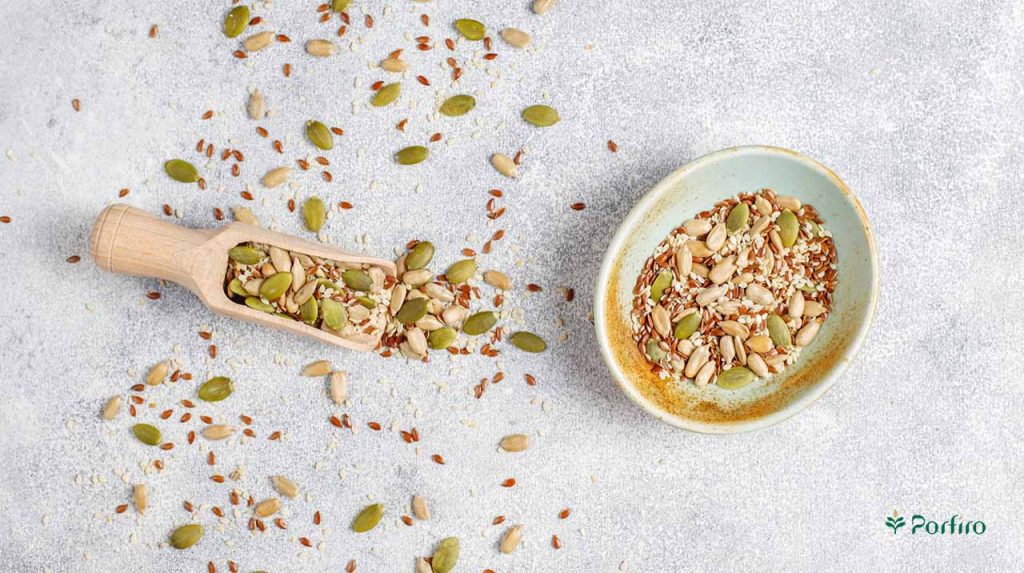 A mixture of different seeds in a bowl, some of which has been poured onto a wooden spoon and some on the floor.