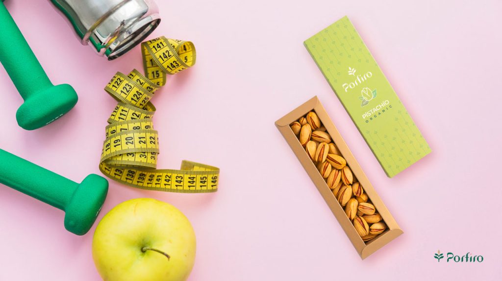 An open package of Porfiro pistachios for weight control next to a scale, measuring tape, and an apple.