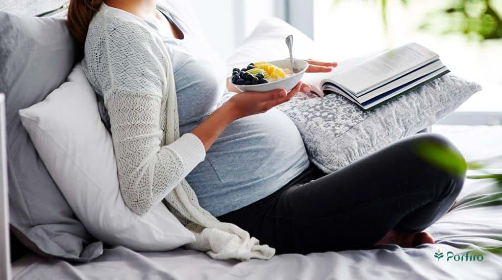 Pregnant woman sitting on her bed with a cushion on her lap, eating a healthy snack while reading
