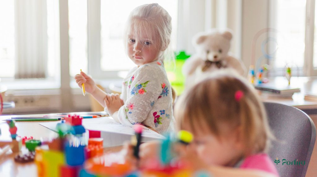 Two children are sitting together in a playroom, one playing and the other looking at her.
