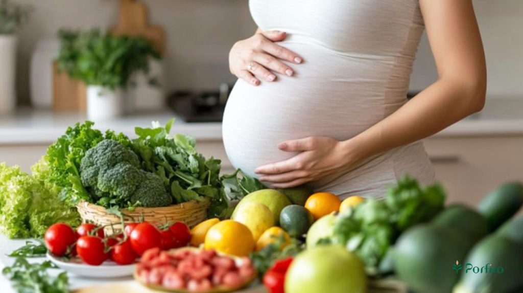 A pregnant woman stands in the kitchen, with a large amount of healthy, fresh fruits and vegetables in front of her. For the article Nutrition in Pregnancy: Essential Nutritional Considerations You Need to Know on the Porfiro blog.